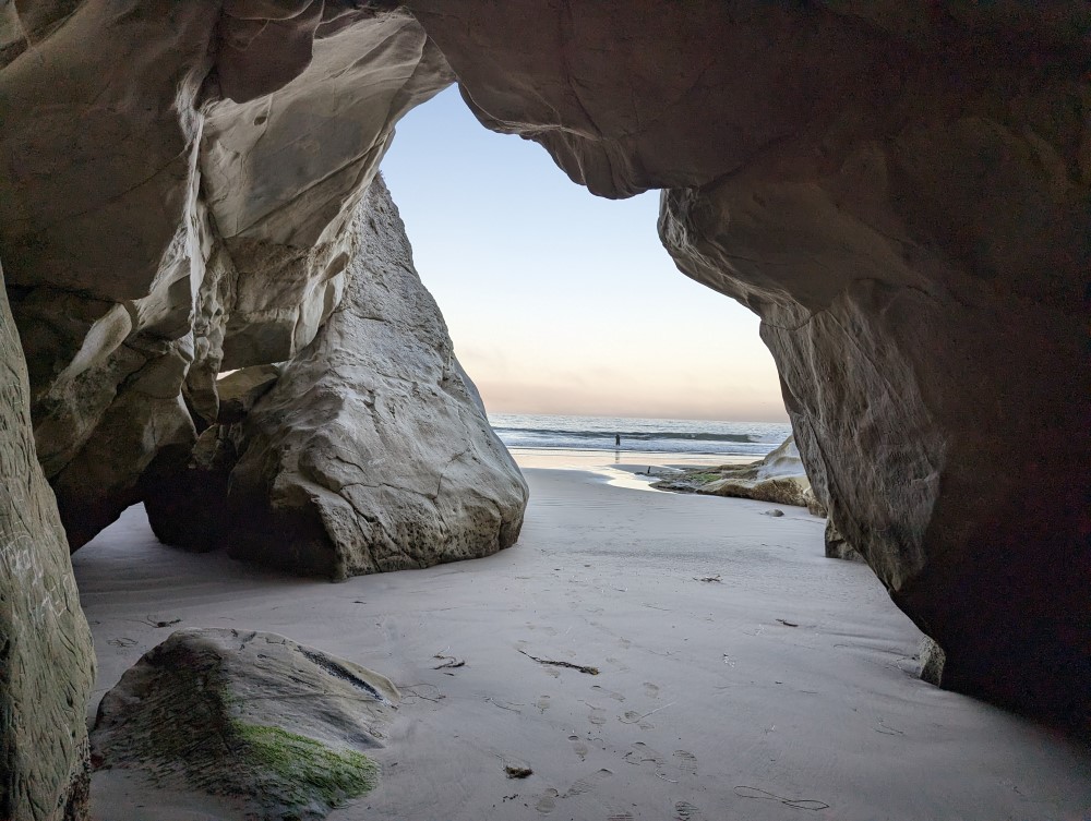Looking out onto a beach from inside a seaside cave. Someone is catching fish, framed by the cave entrance.