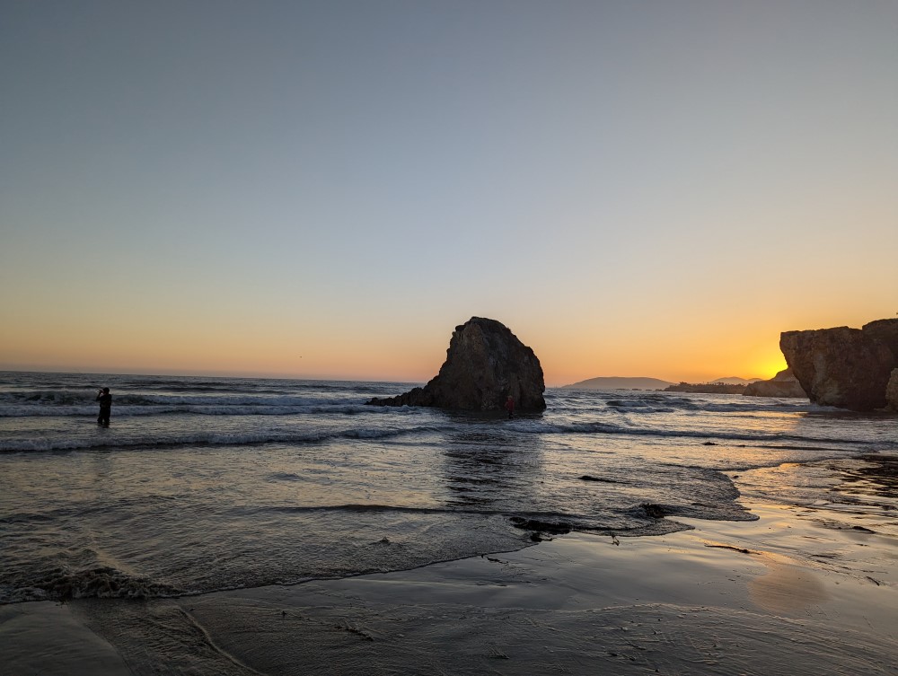 A A beach at sunset. There’s a cliff and a cliff-sized rock next to it, a bit out to sea, and someone catching fish.