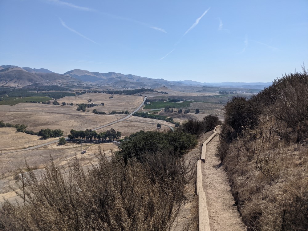 A view from the top of a hill. You can see the path on the side of the hill, a dirt path with a rough wood border, and beyond that a dry valley with patches of green farmland, and beyond that, more hills..